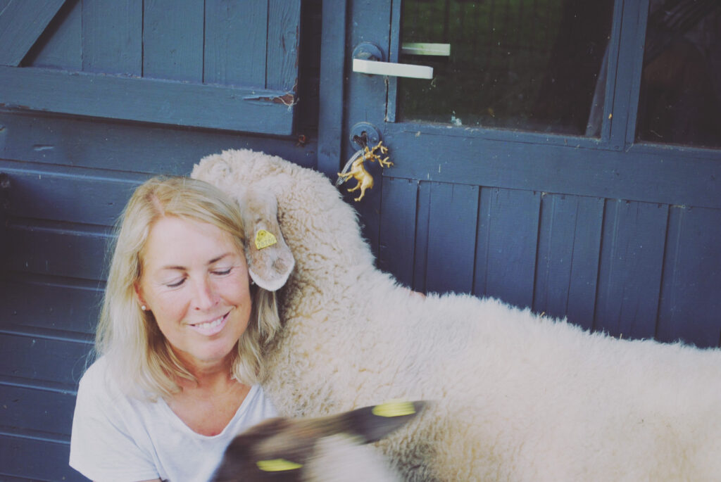 Eine Frau mit blondem Haar sitzt lächelnd neben einem weißen Schaf und streichelt ihren Kopf. Im Vordergrund ist ein weiteres Tier mit einer gelben Ohrmarke teilweise zu sehen. Sie stehen vor einer dunkelblauen Holztür.
