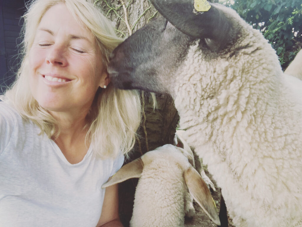 Eine Frau mit blondem Haar und weißem Hemd lächelt mit geschlossenen Augen, während ein Schaf ihren Kopf anstupst. Im Vordergrund ist ein weiteres Schaf teilweise zu sehen. Sie sind draußen, in der Nähe eines Baumes.