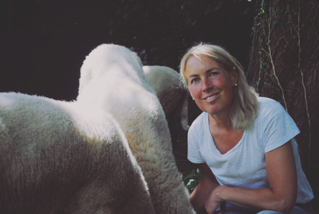 Eine lächelnde Frau mit blonden Haaren und weißem Hemd kniet neben einem Schaf. Im Vordergrund stehen zwei Schafe mit dicker Wolle, von denen eines der Kamera deutlich seinen Rücken zuwendet. Der Hintergrund ist dunkel und bildet einen Kontrast zum hellen Hemd der Frau.