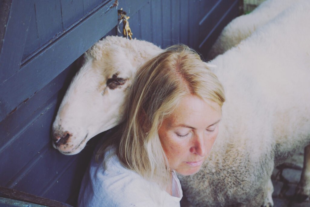 Eine Frau mit blondem Haar und weißem Hemd sitzt neben einem weißen Schaf. Das Schaf lehnt seinen Kopf an der Schulter der Frau und beide wirken ruhig und gelassen. Sie stehen neben einer blauen Holzkonstruktion.